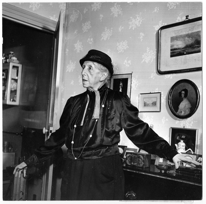 Schwarz-Weiß-Aufnahme einer Frau (Anette Kolb) mit Hut und ausgebreiteten Armen in Wohnzimmer