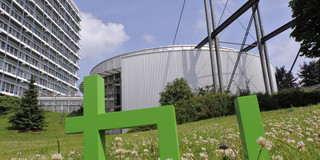 Bild des Logos der TU Dortmund platziert auf einer Wiese zwischen Gebäuden der Universität.