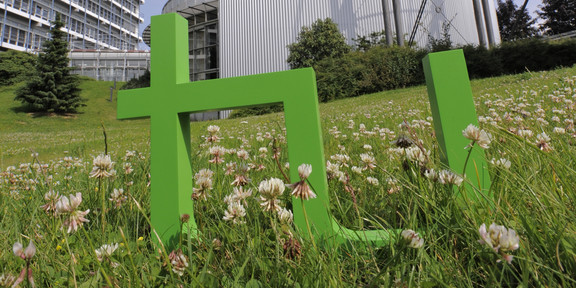 Bild des Logos der TU Dortmund platziert auf einer Wiese zwischen Gebäuden der Universität.