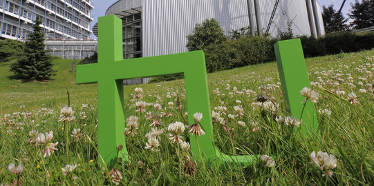 Bild des Logos der TU Dortmund platziert auf einer Wiese zwischen Gebäuden der Universität.